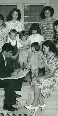 Black and white photograph showing a census taker sitting on the front steps of a house with  a woman and many children who are observing him record their answers
