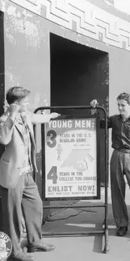 Two young men stand outside a recruiting office. Between them is a poster discussing the benefits of enlisting in the Army.