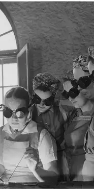 Black and white photograph showing a number of women wearing protective goggles while watching a demonstration of an industrial task.