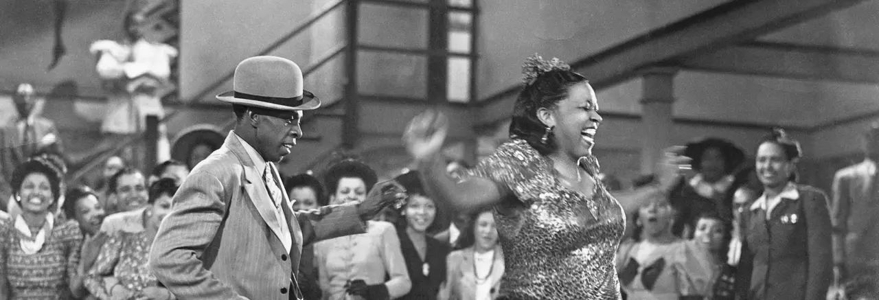 A  Black man in a light-colored jacket, white shirt, light-colored patterned tie, and light-colored hat with a dark brim dances next to a smiling Black woman wearing a dark, sparkly dress.