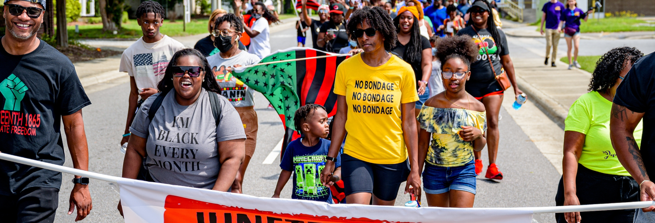 A color photograph of a parade in celebration of Juneteenth; men, women, and children predominantly wearing T-shirts and shorts marching on a city street; in foreground, two Black men wearing black Juneteenth t-shirts smile as they hold a banner reading "Juneteenth: Celebrate Freedom June 19th."