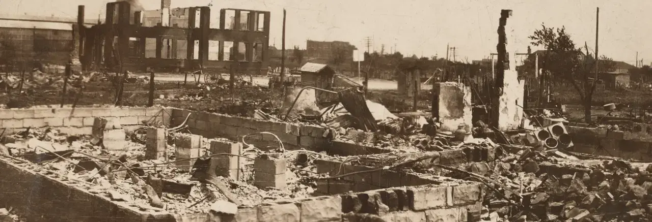 A black-and-white photograph of the destroyed Greenwood District in Tulsa, Oklahoma after the Tulsa Race Massacre. In the foreground is a stone foundation littered with and surrounded by ash. The background features remnants of building façades.