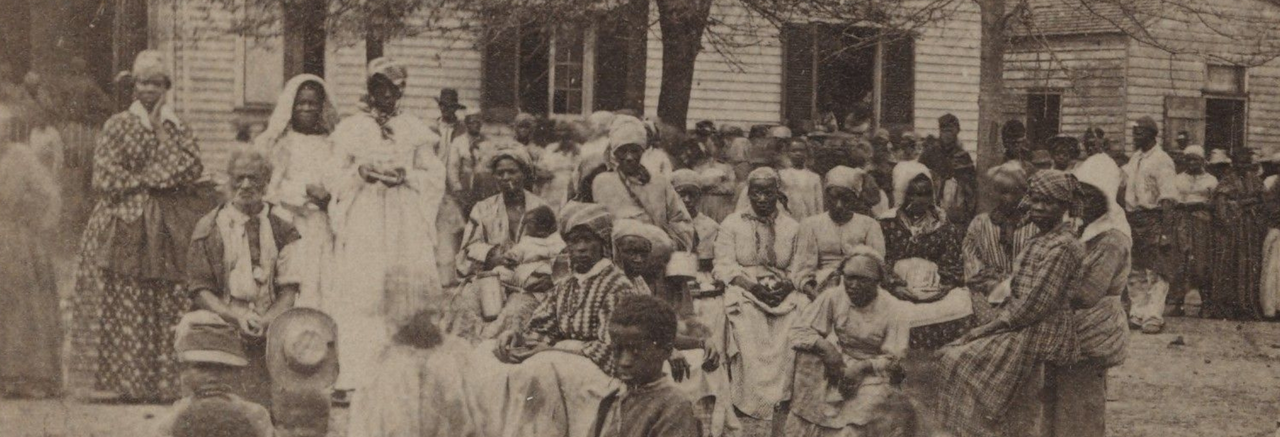 Black men, women, and children sitting and standing in a field in front of two trees and two houses.