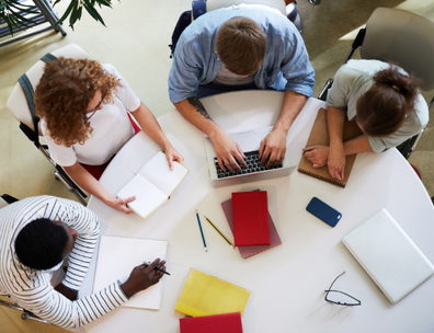 View from above showing four students gathered around a round table with notepads and laptops