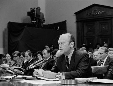 Gerald Ford seated in front of a microphone reading from a piece of paper in a House of Representatives hearing room, as others listen in among the gallery. In the background is a television camera and camera operator.