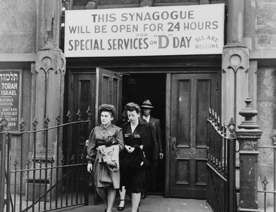 Two women in dark hats and dresses walking out of a synagogue. A man in a dark suit, white shirt, and light-colored hat is behind the women. Above the doorway of the synagogue is a sign saying "This synagogue will be open for 24 hours for special services on D-Day: All Are Welcome"