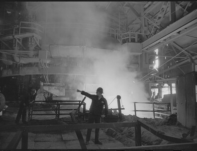 Black and white photograph of a steelworker pointing to another, with steam rising behind him. 