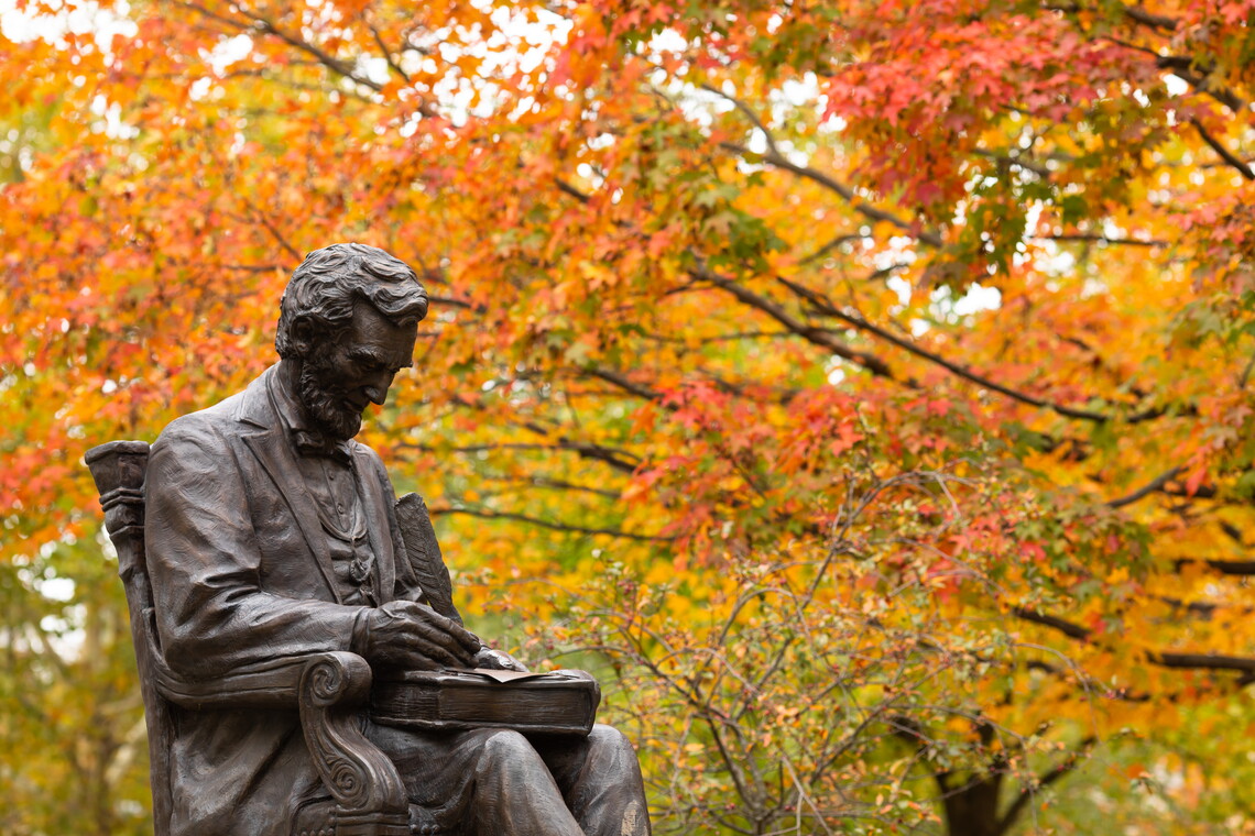Photograph taken during autumn, showing statue of Lincoln with quill in hand, on Gettysburg campus, fall folliage in background