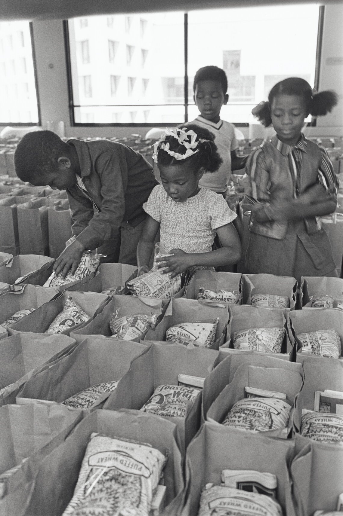 A black-and-white photograph showing children preparing bags of food to be distributed through the Panther Free Food Program at the Black Panther Community Survival Conference in Oakland, California. Four Black children, two boys and two girls, are placing bags of puffed wheat cereal into brown paper grocery bags that have been organized in rows.