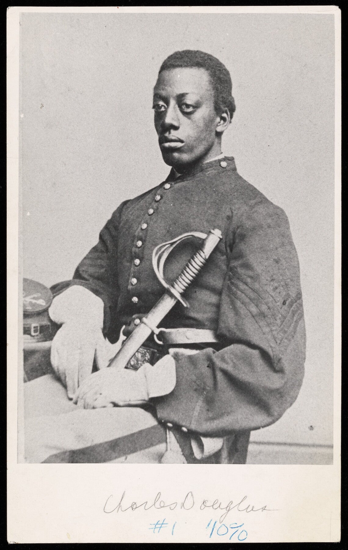 A black-and-white photograph of a serious young Black man in a military uniform (dark coat, white gloves, light-colored pants with a dark racing stripe) holding a sheathed sword