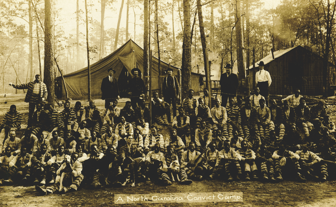 Image 1 (obverse): A sepia photograph of several dozen men (mostly Black men) in striped prison jumpsuits sitting around some trees, with a tent and a cabin behind them. Several of the men have dogs sitting with them. One man holds a guitar. Standing behind the prisoners are five White men in suits. White text in a lighter colored rectangle at the bottom reads "A North Carolina Convict Camp."