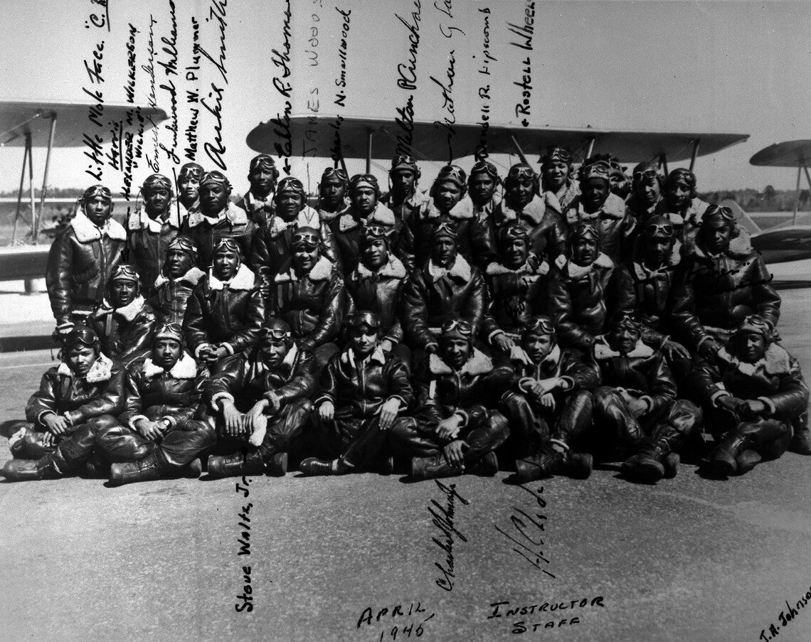 Several Black men in three rows (sitting, kneeling, standing), all in dark flight suits, boots, and goggled helmets, in front of biplanes. Some men's signatures are next to their photograph.