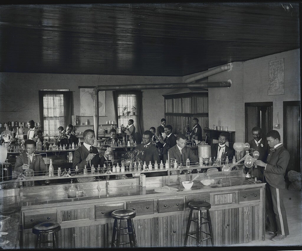 A photograph of a chemistry laboratory, with three lab benches in the foreground, midground, and background. Black men in white shirts, dark coats, and dark ties are working at each bench. In the right foreground, framed by a doorway behind him, is George Washington Carver, a mustachioed Black man wearing a dark coat and tie, looking down at another chemist's work.