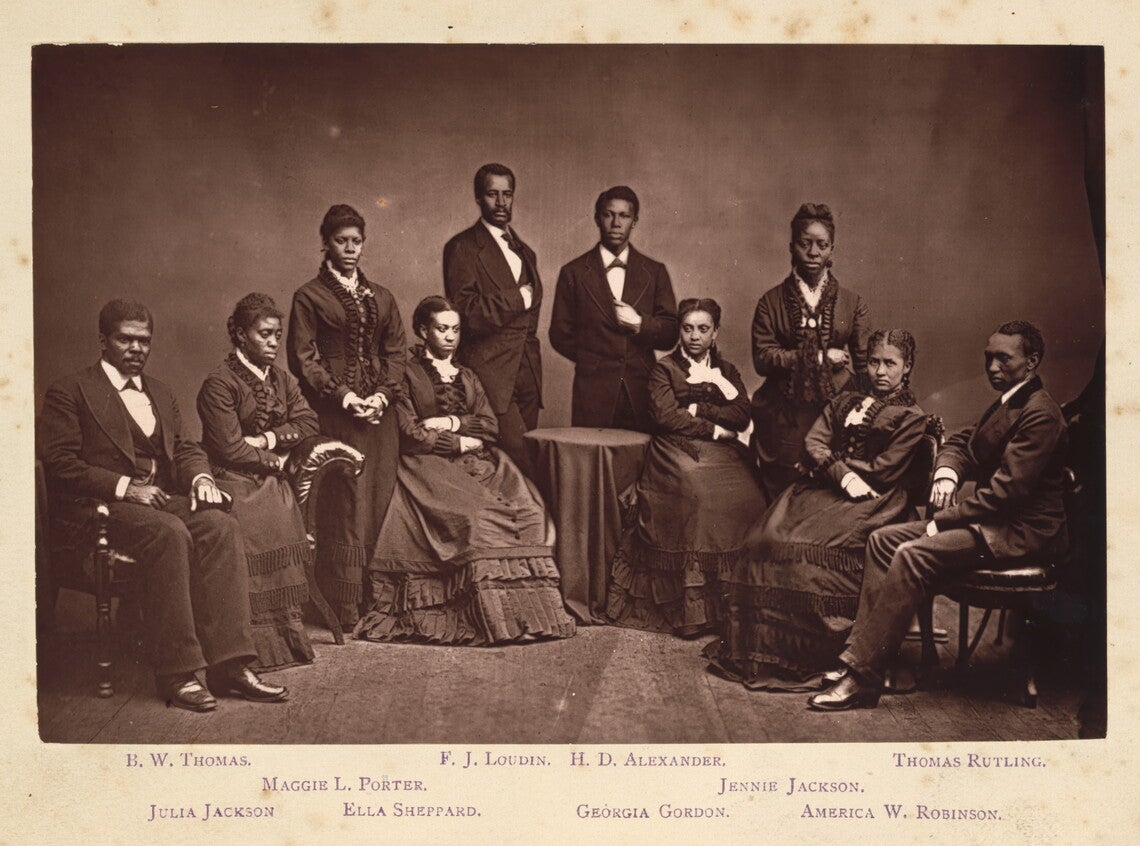Ten Black men and women sitting and standing while wearing dark suits or dresses. A small table with a dark tablecloth sits in the middle background.