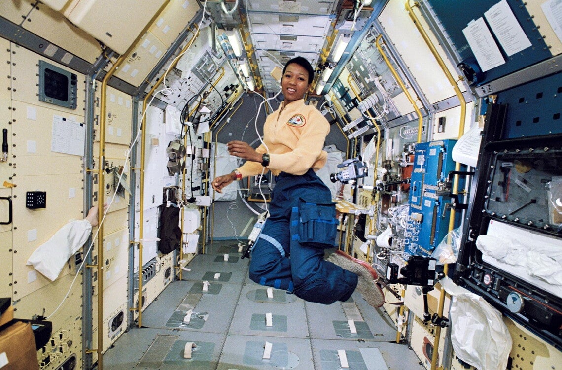 A color photograph of Mae Jemison, a Black woman, wearing an astronaut's uniform (yellow shirt with mission patch, blue cargo pants, grey slippers) aboard the space shuttle. She is floating in zero gravity, surrounded by science stations.