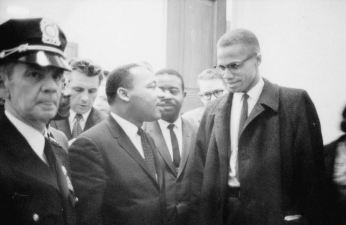 A black-and-white photograph of two Black men, Martin Luther King Jr. and Malcolm X, looking at one another as other Black and White men look on. A White police officer in uniform stands to the left.