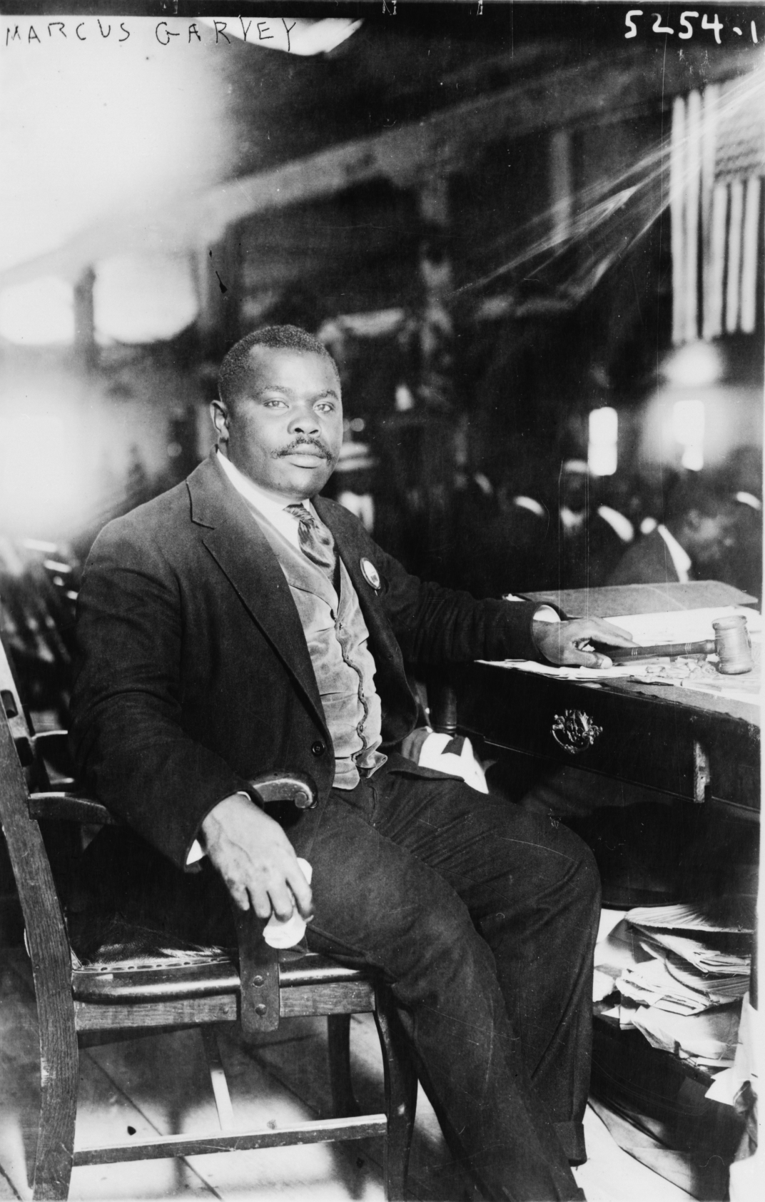 A black-and-white photograph of Marcus Garvey, a Black man wearing a dark suit, light-colored vest, white shirt, and dark tie, sitting in a wooden chair next to a desk filled with papers, holding a handkerchief in his right hand and a gavel in his left.