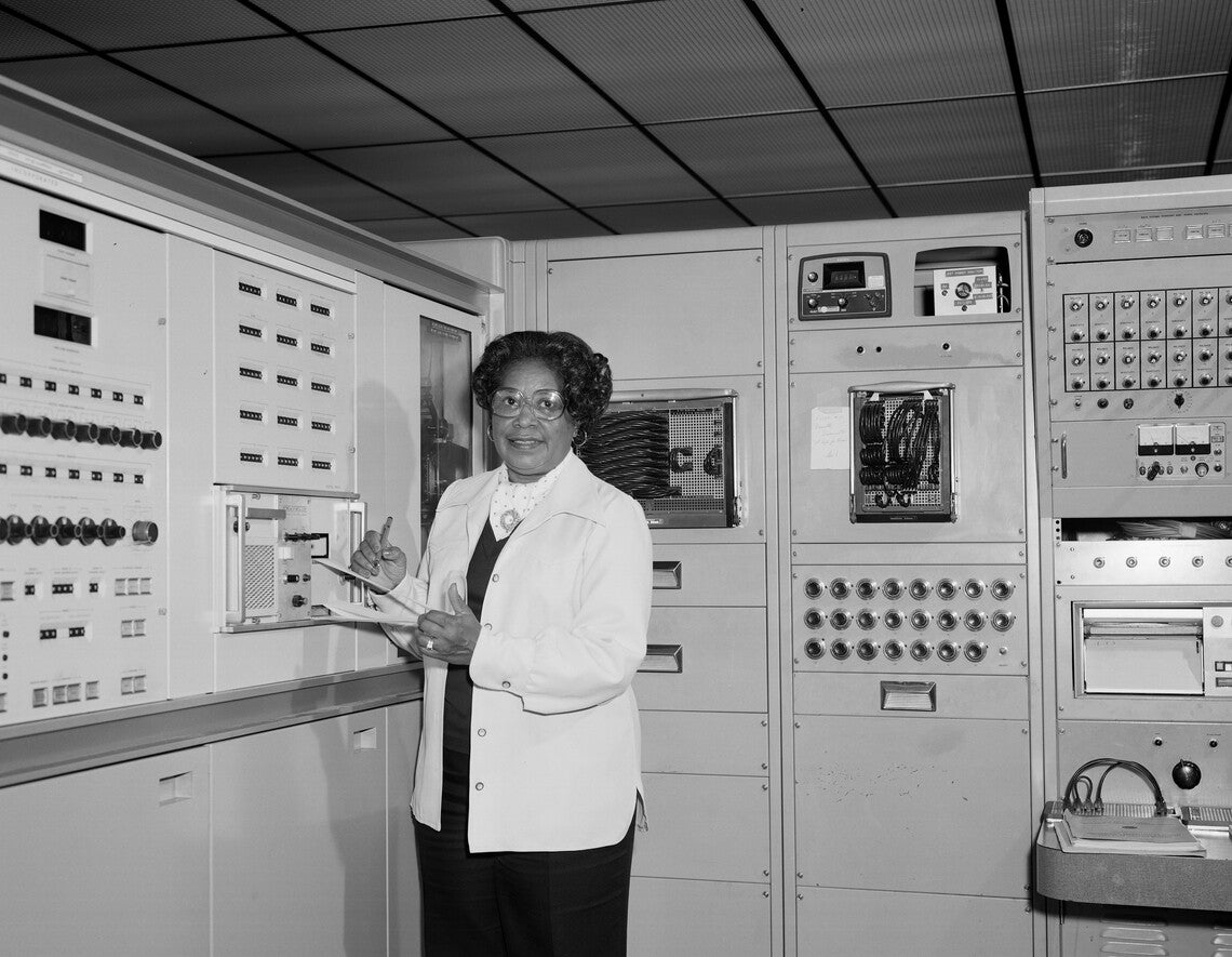 A black-and-white photograph of Mary Jackson, a Black woman wearing glasses and a white lab coat, writing on a clipboard and surrounded by computer equipment