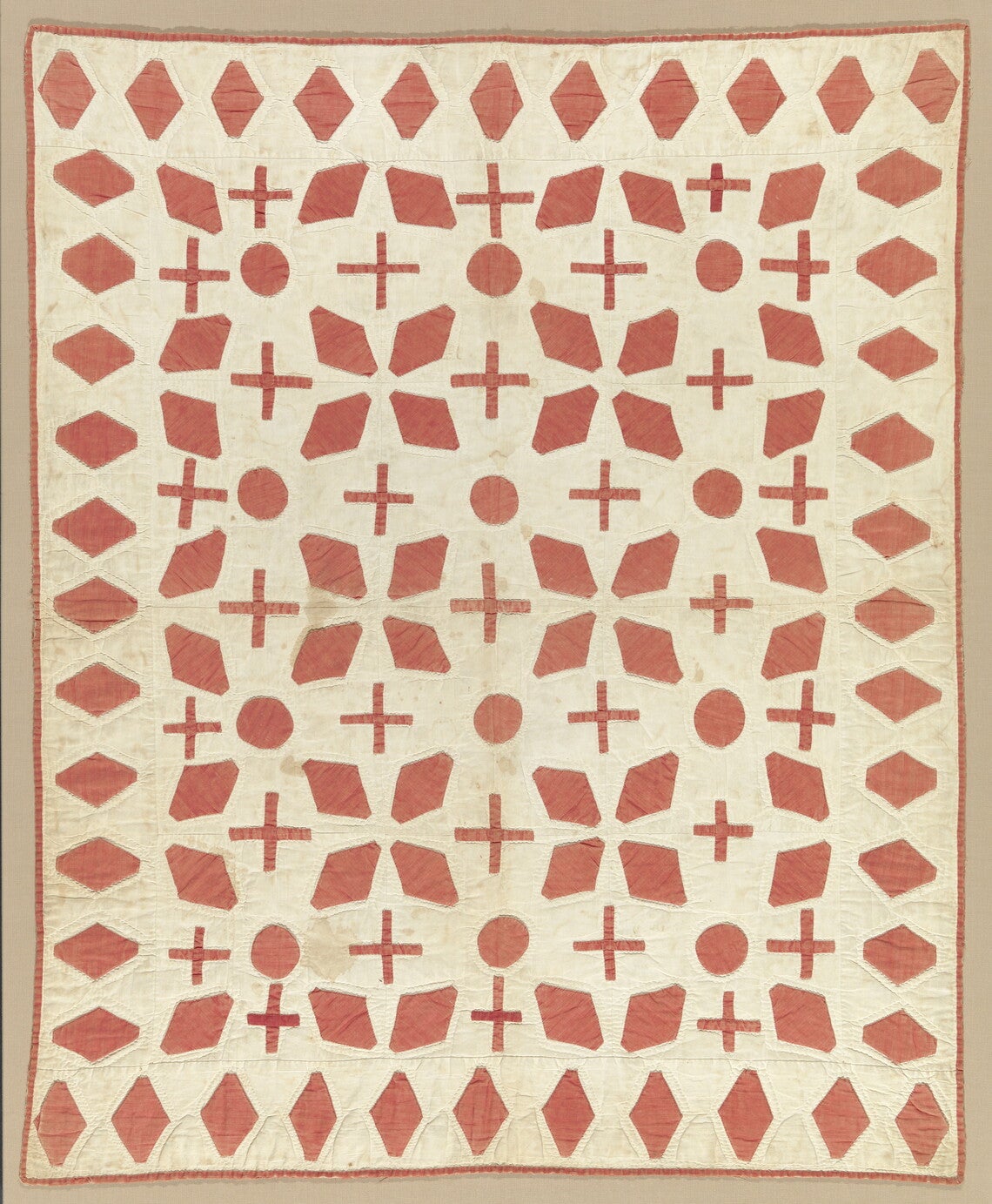 A cream quilted bedcover with appliqued red diamonds, circles, and crosses and edges bound with red