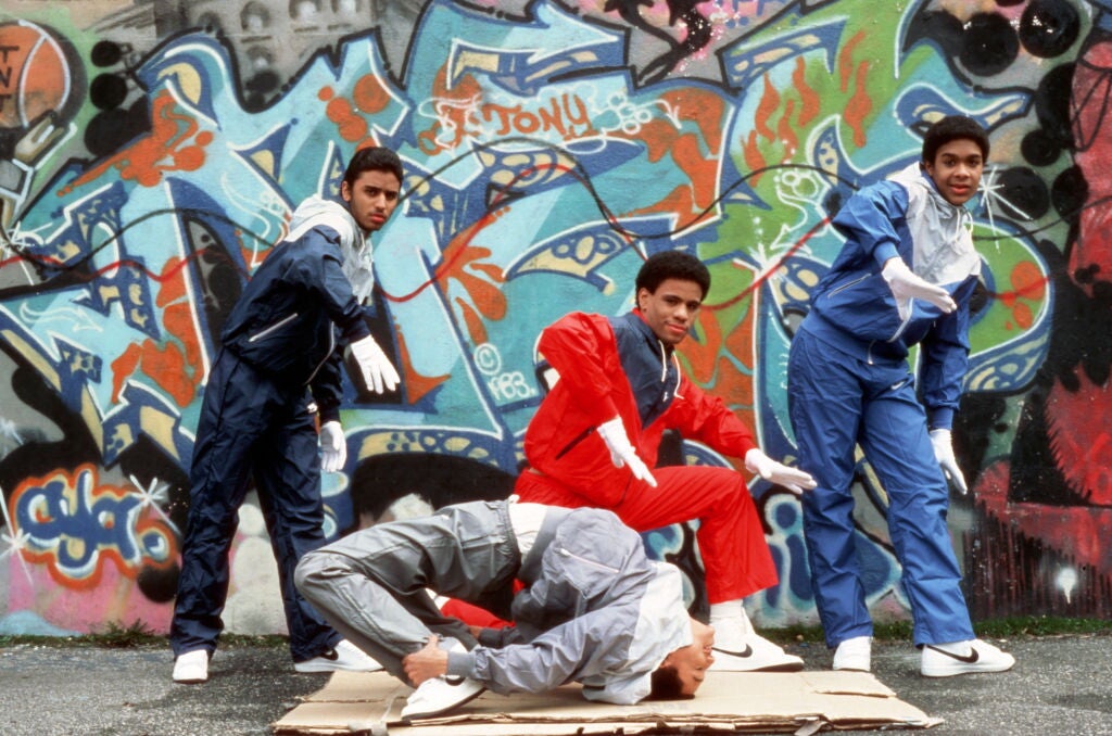 Three young Black men wearing tracksuits and white gloves dancing as another young Black man inverts his body on a piece of cardboard. Behind them is a graffitied wall.