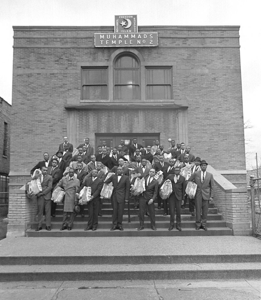 A black-and-white photograph of about forty Black men in dark suits and ties holding bundled stacks of newspapers under their arms, standing on the steps of a brick building topped with a crescent and the words "Muhammad's Temple No. 2."
