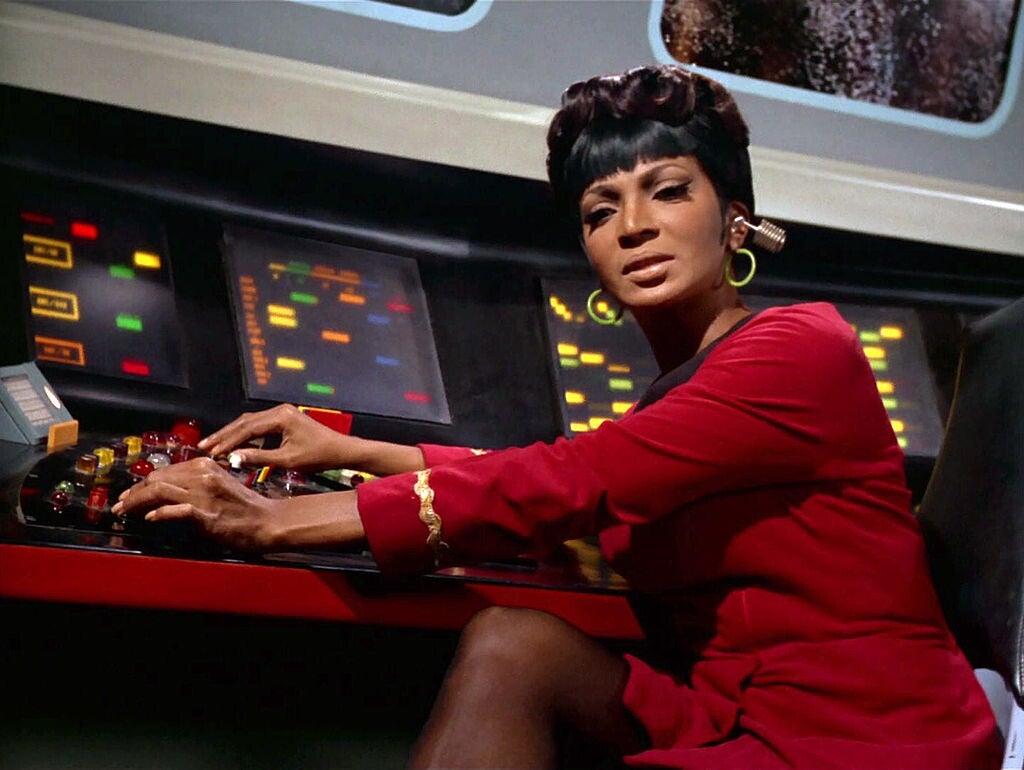 Lieutenant Uhura (Nichelle Nichols), a Black woman, wearing a Starfleet uniform (red dress with a black collar and a wavy gold band on each sleeve), with a communications earpiece in her left ear and green hoop earrings. She has her hands on a bank of buttons connected to an array of four light screens.