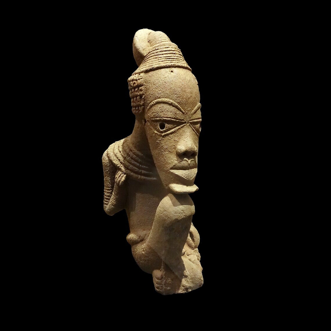 A stylized sculpture of a seated man with an oversized and elongated head relative to their body, chin resting on the figure's knees. Damage to the figures shins.