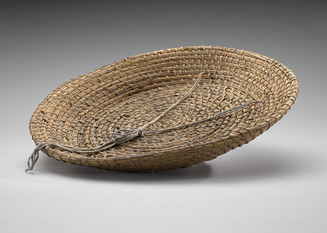 A large shallow, round tray shaped basket made of reeds, rush and palmetto coiled and woven together. A faded purple string is woven through the basket on one side and knotted inside the basket.