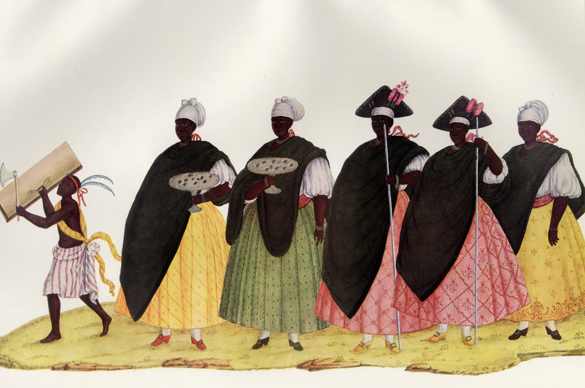 A group of five Black women wearing white and black headdresses, black frocks, and brightly colored skirts, follows a small boy wearing colorful clothing adorned by feathers, holding a piece of wood and a small ax.