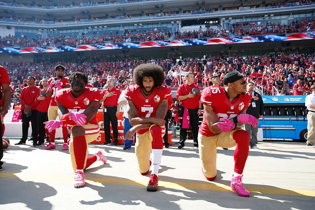 Three of the San Francisco 49ers football team kneeling (right knee on ground, arms balanced on left knee) during the pregame performance of the national anthem, as their teammates, coaches, and staff stand, some with hats or hands on their left breast, behind them. The three Black men kneeling are wearing the 49ers uniform of red jerseys, gold pants (with red-white-red stripes along the leg), and either red or white socks.