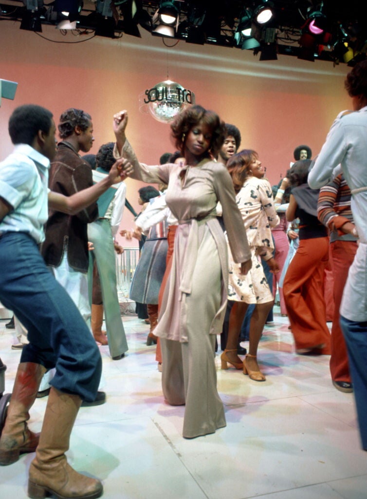 A Black woman wearing a beige jumpsuit with a cinched waist dancing under a mirrorball reading "Soul Train," in the middle of a crowd of other Black men and women dancing.