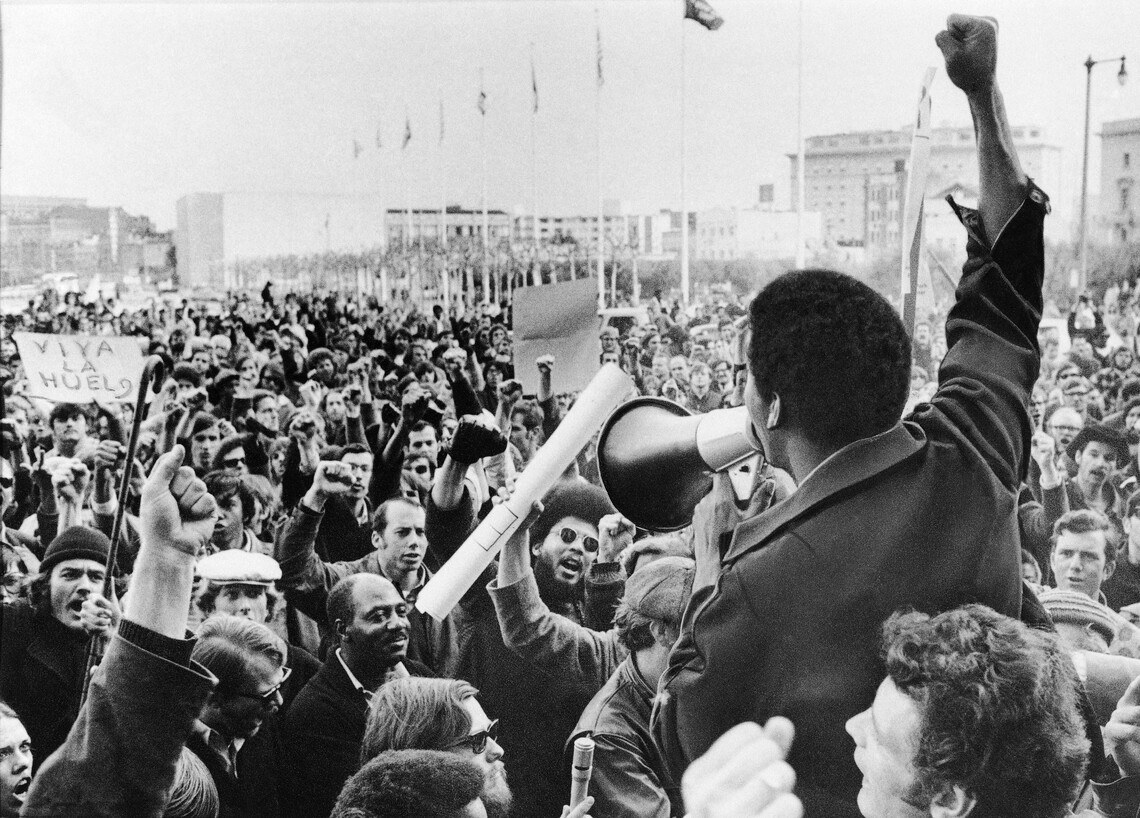 A Black man wearing a dark coat speaking into a bullhorn megaphone held in his left hand, while his right arm is outstretched with a clenched fist. A crowd looks on and raises their right fists in the air. There is a sign reading "Viva La Huelga" in the left midground.