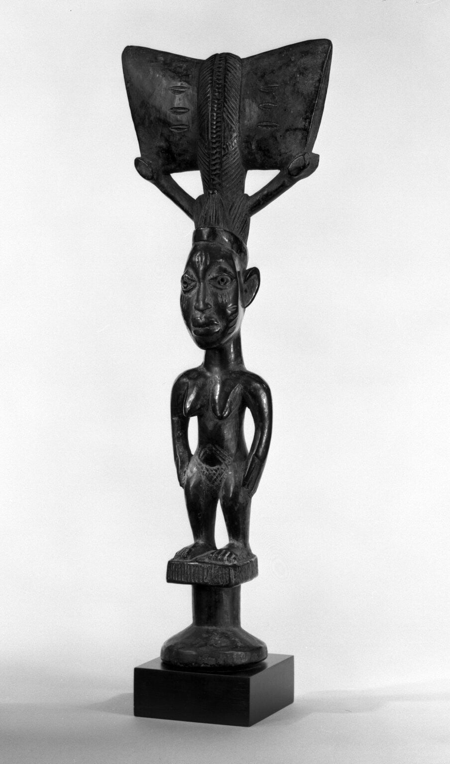 A dance wand made of wood painted black in the form of a standing wooden female figure with arms at sides apart from torso; hands joined at hips; and feet and legs apart.