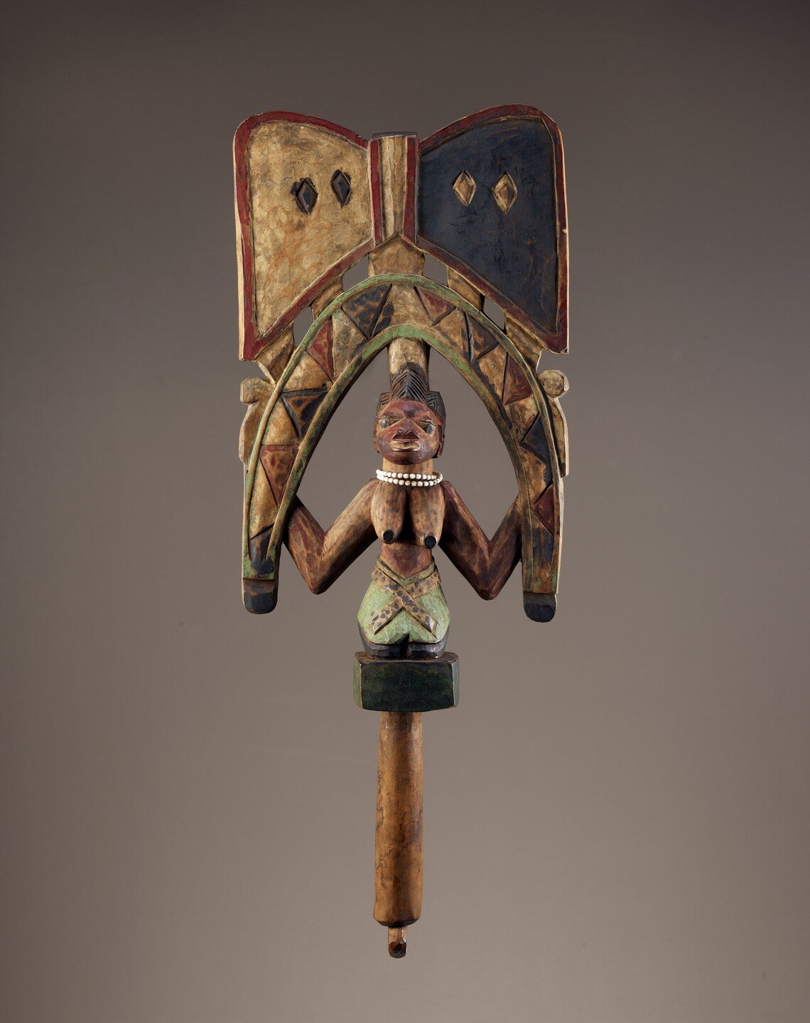 Wood staff in the form of a kneeling female figure holding a stylized snake in an inverted U over her head, topped by a pair of opposing triangles in a bowtie form. Female figure has white beads around her neck and staff is pigmented in red, blue and white.