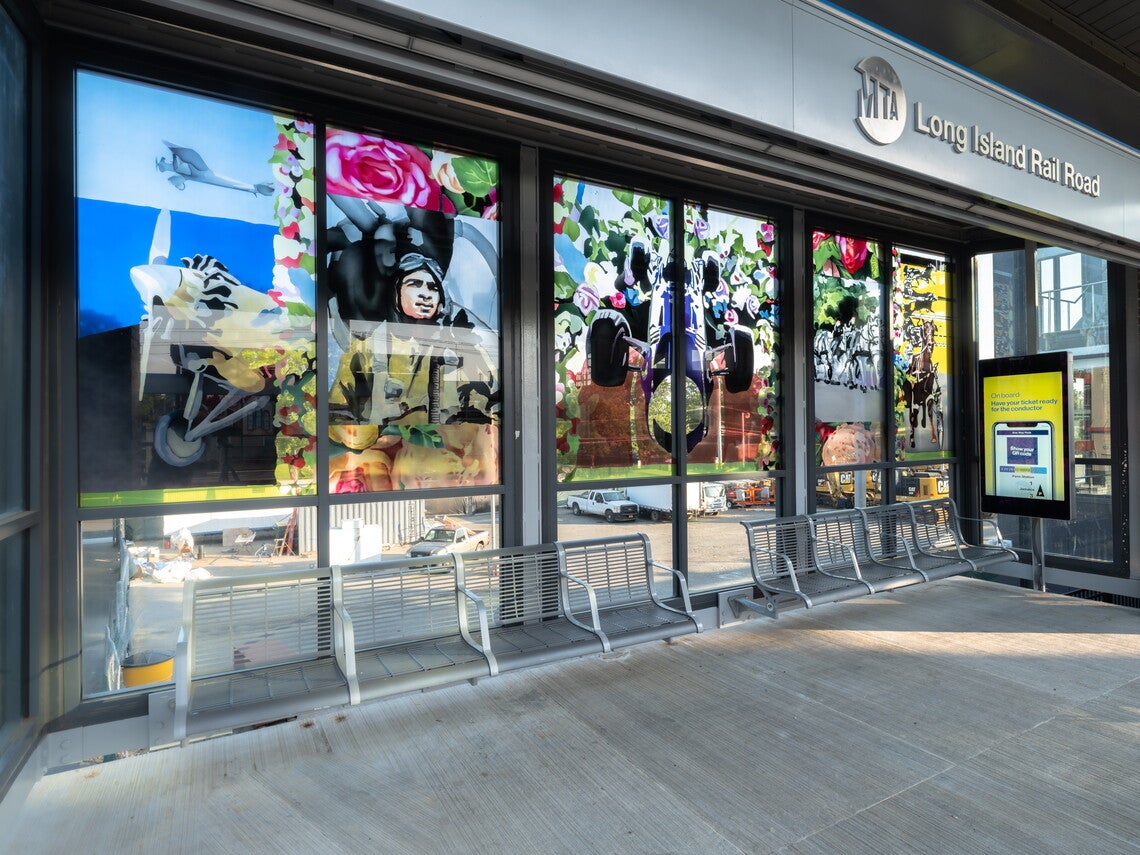 A color photograph of an outdoor train platform with a wall of glass panels overlooking the street below; the images include a propeller airplane, a Tuskegee airman, a car, and horse racing
