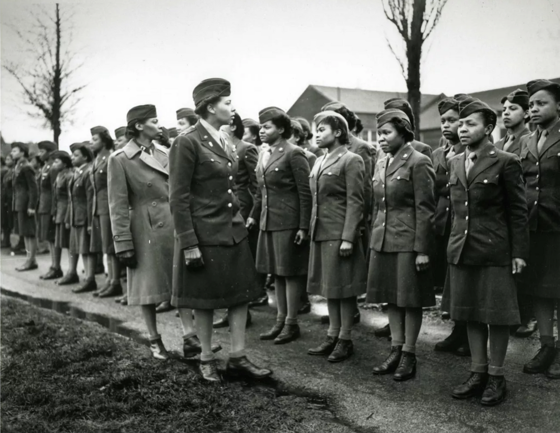 Two Black women in military uniform (dark jackets, light shirts and ties, dark skirts and boots) looking on at two rows of other Black women in military uniform standing at attention.