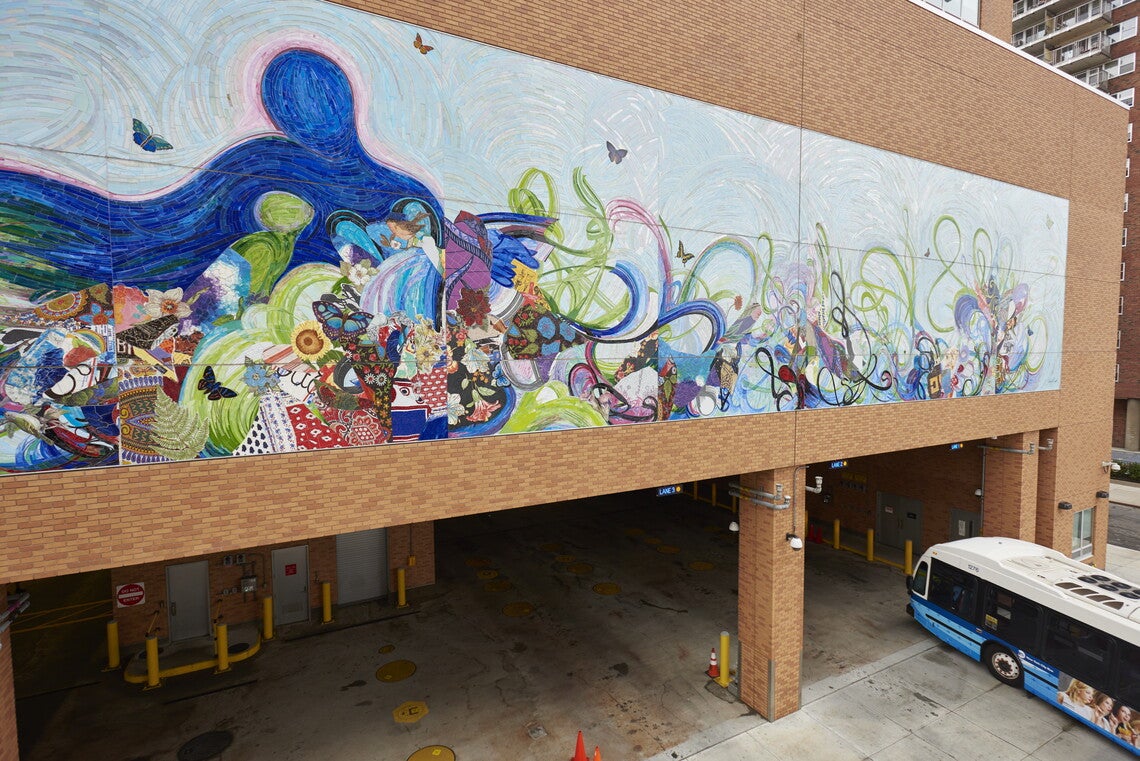 An abstract multicolored mural above a bus depot, with a blue and white bus pulling into the depot in the right foreground