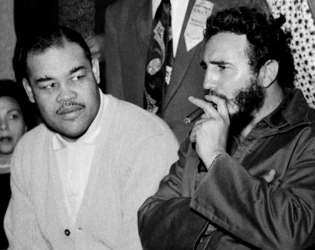 Joe Louis, a Black man wearing a light colored cardigan sweater and white polo shirt, next to Fidel Castro, a bearded Latino man wearing a dark coat and smoking a cigar held in his left hand.