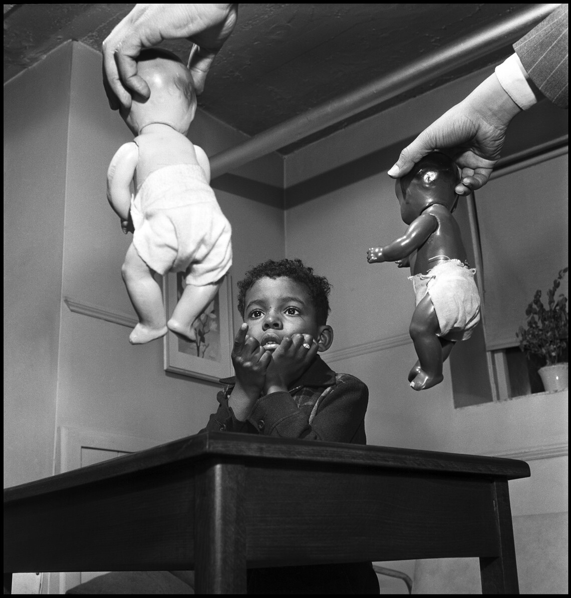A young Black boy sits, heads in hands, and points toward a white baby doll held in the left hand of Kenneth Clark, a Black man whose white shirt and gray pinstriped jacket sleeves are visible. in Dr. Clark's right hand is a black baby doll.