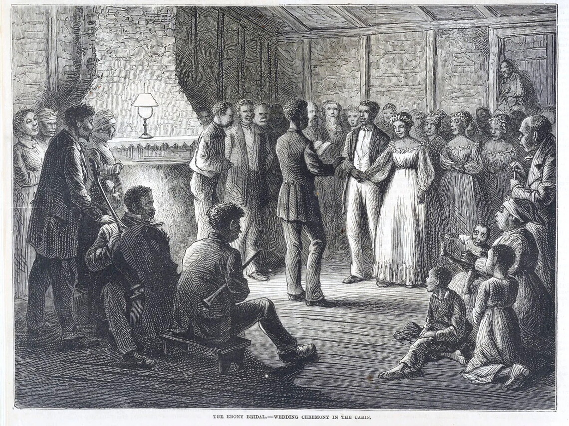 A wedding near a fireplace. The groom is a Black man wearing a dark coat, white pants, and a white shirt. The bride is a Black woman wearing a white dress. Groomsmen and bridesmaids are behind the couple, and the officiant (wearing a dark coat and pants and holding a book) is faced in the couple's direction. Behind the officiant are more witnesses, a bassist, and a clarinetist (the latter musicians sitting).