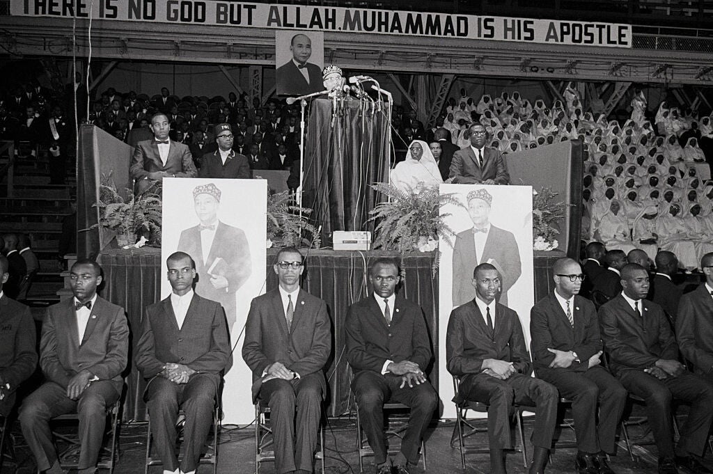 A black-and-white photograph of Elijah Muhammad, a Black man wearing a kufi, barely visible above a lectern and a bank of microphones on a stage. He is flanked on his stage by two Black men on his right and a Black woman and Black man on his left. Behind the stage are rows of Black men in dark suits and ties and Black women in white robes and head coverings in stadium-style seating. Seven black men in dark suits are seated looking out into the audience. Two large posters of Elijah Muhammad are posted in fro