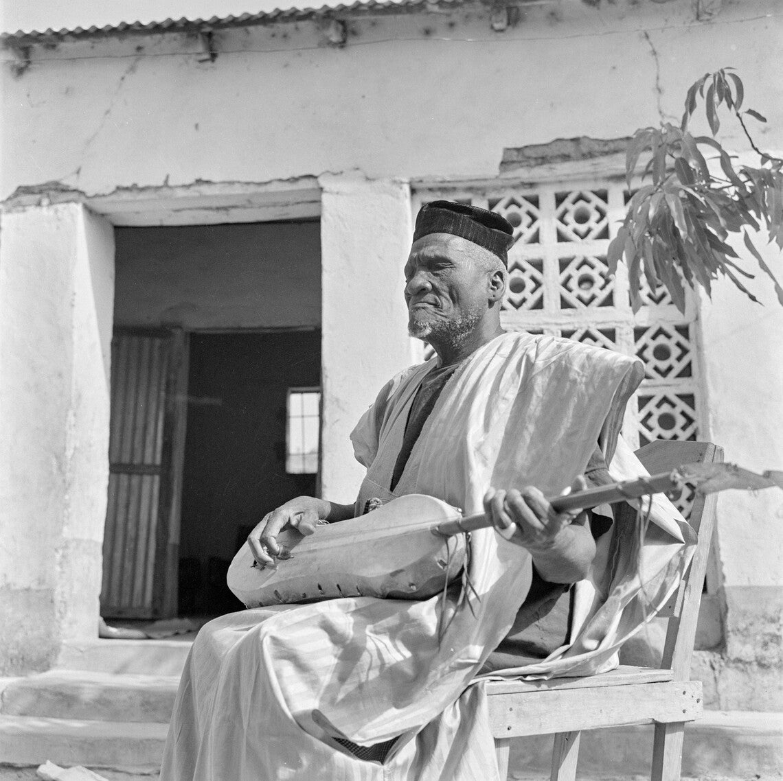 A goateed Black man in a white robe and black kufi hat sitting with eyes closed, plucking a guitar made of a calabash squash.