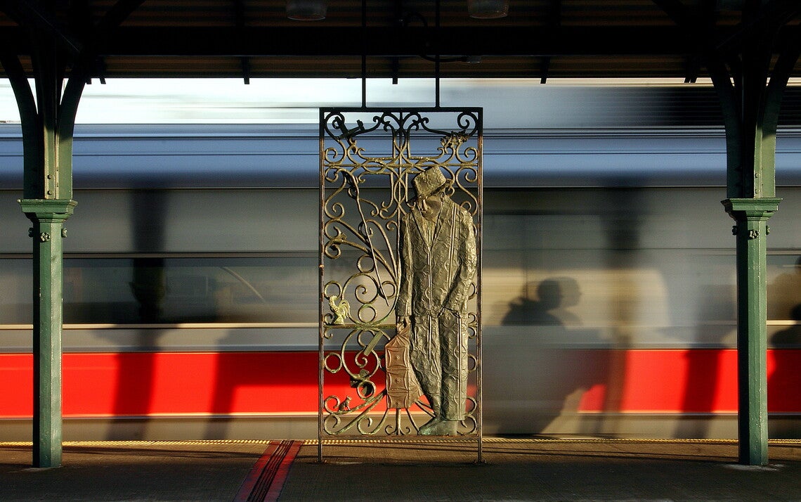 Hear...: A color photograph of an outdoor train platform with, at center, a wrought-iron sculpture of a man in a suit and hat holding a suitcase.