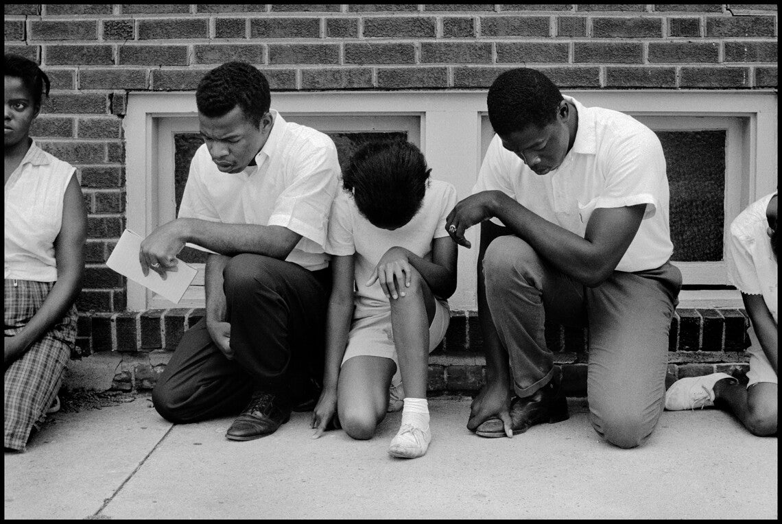 A black-and-white photograph of four African Americans kneeling in prayer on a sidewalk in front of a brick building. From left to right: a Black woman wearing a white sleeveless blouse and plaid shorts kneeling with head raised, looking straight ahead; John Lewis, a Black man wearing a white button-down shirt, black pants, and black shoes, kneeling with head bowed, holding a paper in his left hand; a Black girl wearing a white shirt, shorts, and white shoes, kneeling with head bowed; and a Black man wearin