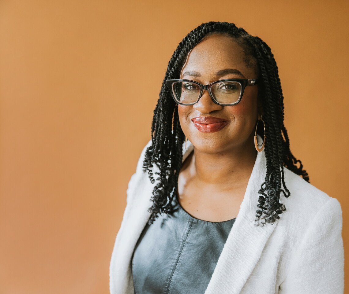 Photograph of Prof. Keisha Blain, a Black professor, smiling while wearing a white jacket over a textured green shirt, black glasses, and gold earrings.