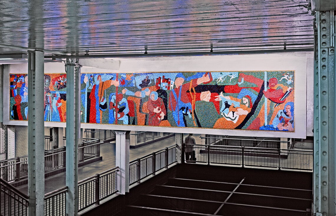 A color photograph of an impressionist, multicolored mosaic mural of people on a subway train in the background, with beams holding up the ceiling of a subway station in the foreground.