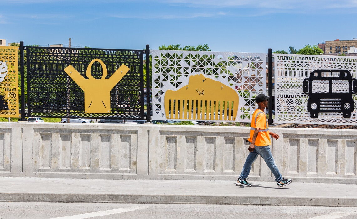 A color photograph of an overpass, the wall of the overpass consists of a concrete wall topped by perforated metal panels, through which train tracks and a parking lot are visible. The metal panels are white, black, and yellow. A Black man wearing a black baseball cap, orange shirt, jeans, and sneakers, is walking along the overpass.