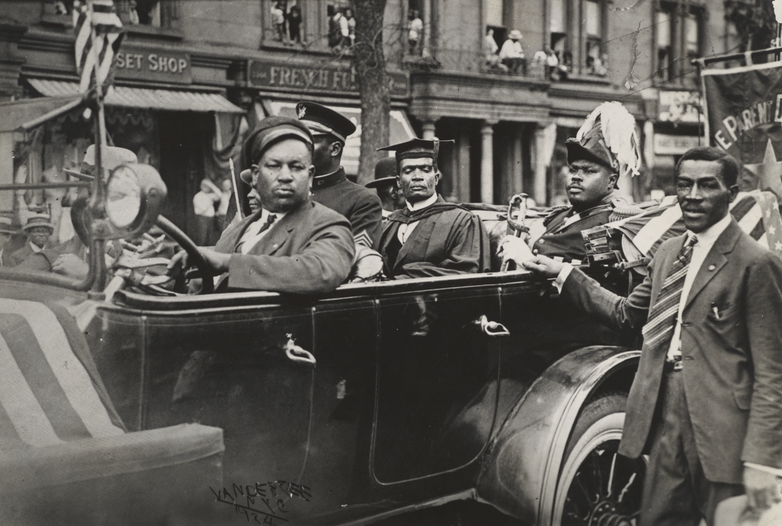 An open-topped car with four passengers in a parade. The driver is a Black man with a dark jacket, white shirt, dark tie, and dark driver's cap. In the back seat, from left to right, is a Black man in a military uniform (dark jacket, dark cap with gold flourishes); a Black man wearing academic regalia (dark robe and hood, dark mortarboard and tassel); Marcus Garvey, a Black man, wearing a military uniform (dark jacket, dark hat with a white feather) and holding a sword. Next to the car is a Black man wearin
