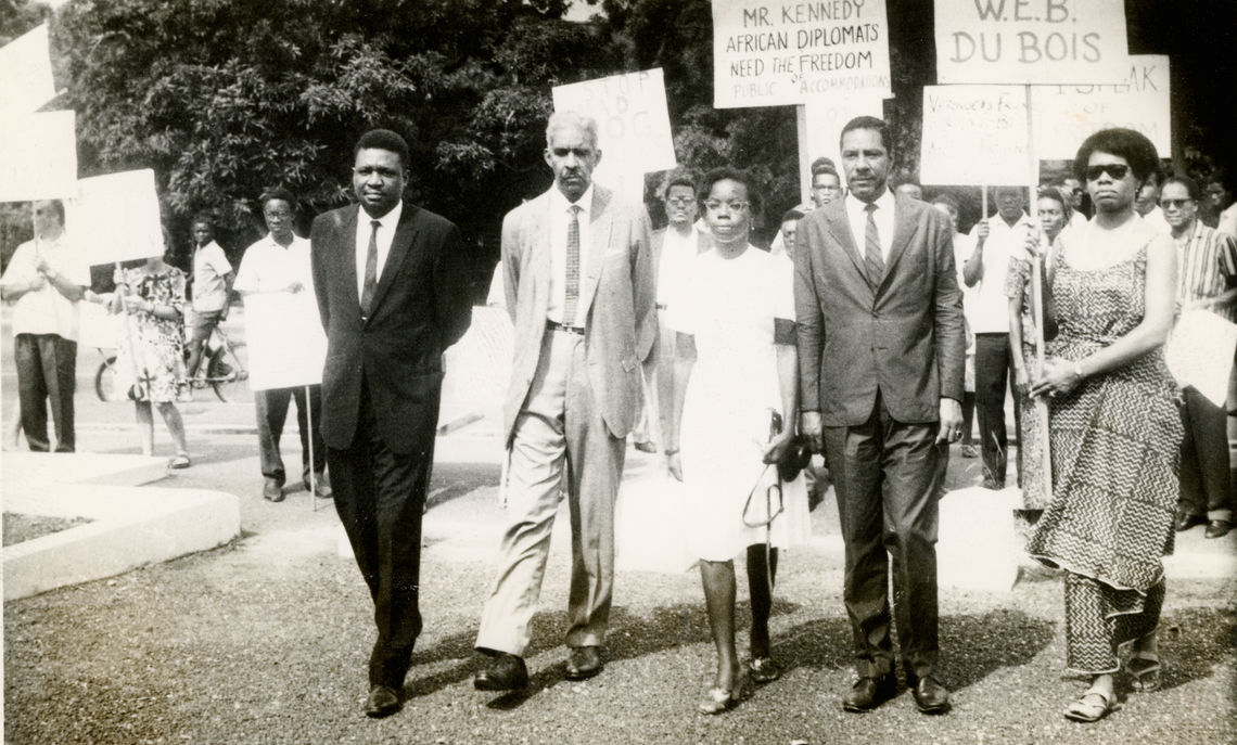 Three Black men in suits and two Black women in dresses protesting, as other people protest in the background. The woman on the right, Maya Angelou, is holding a sign, the bottom of which reads "W.E.B. Du Bois."