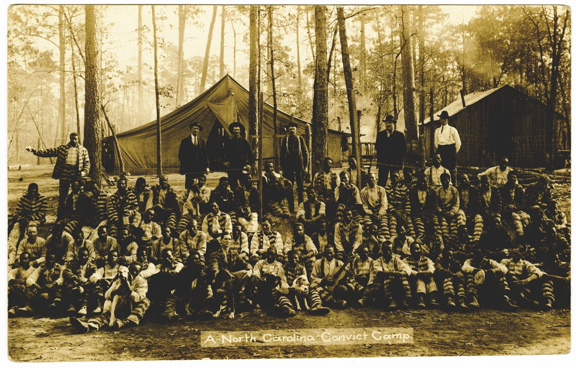 A sepia photograph of several dozen men (mostly Black men) in striped prison jumpsuits sitting around some trees, with a tent and a cabin behind them. Several of the men have dogs sitting with them. One man holds a guitar. Standing behind the prisoners are five White men in suits. White text in a lighter colored rectangle at the bottom reads "A North Carolina Convict Camp."